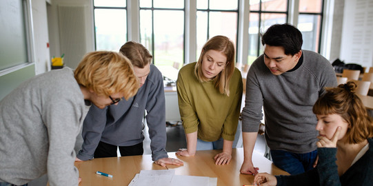 Studierende Gruppenarbeit Fünf Studierende im Seminarraum stehen um einen Tisch herum und sehen auf ein Dokument