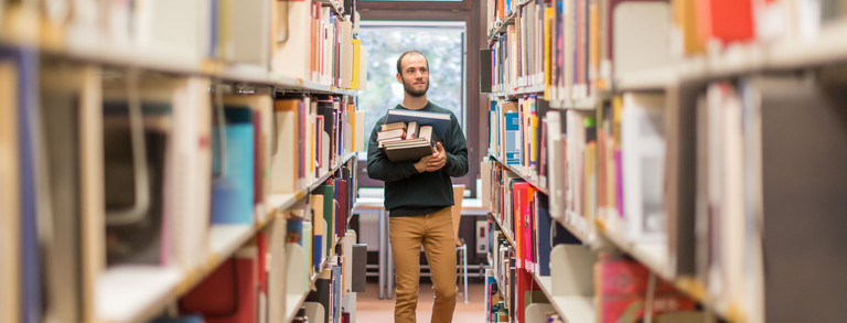 Studierender läuft an den Bücherregalen in der Bibliothek entlang.