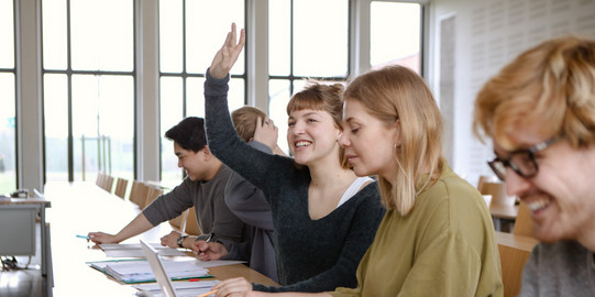 Studierende im Seminar Fünf Studierende sitzen in einem Seminarraum an Tischen. Eine Studentin hebt ihre Hand.