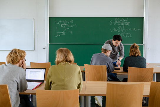 Vier Studierende im Seminarraum sitzen und arbeiten zusammen mit dem Dozierenden im intergrund.